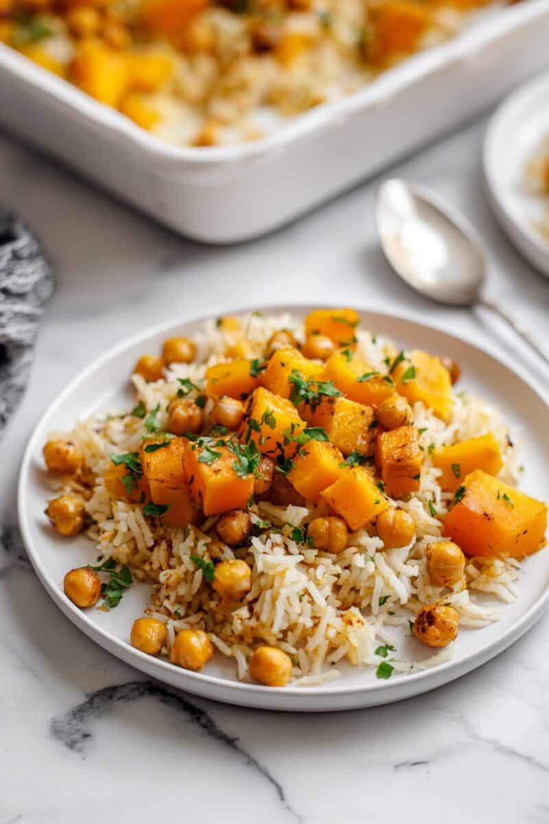 A serving of vegan roasted butternut squash and chickpea rice bake on a white plate, topped with fresh parsley, highlighting the golden roasted vegetables and fluffy rice.