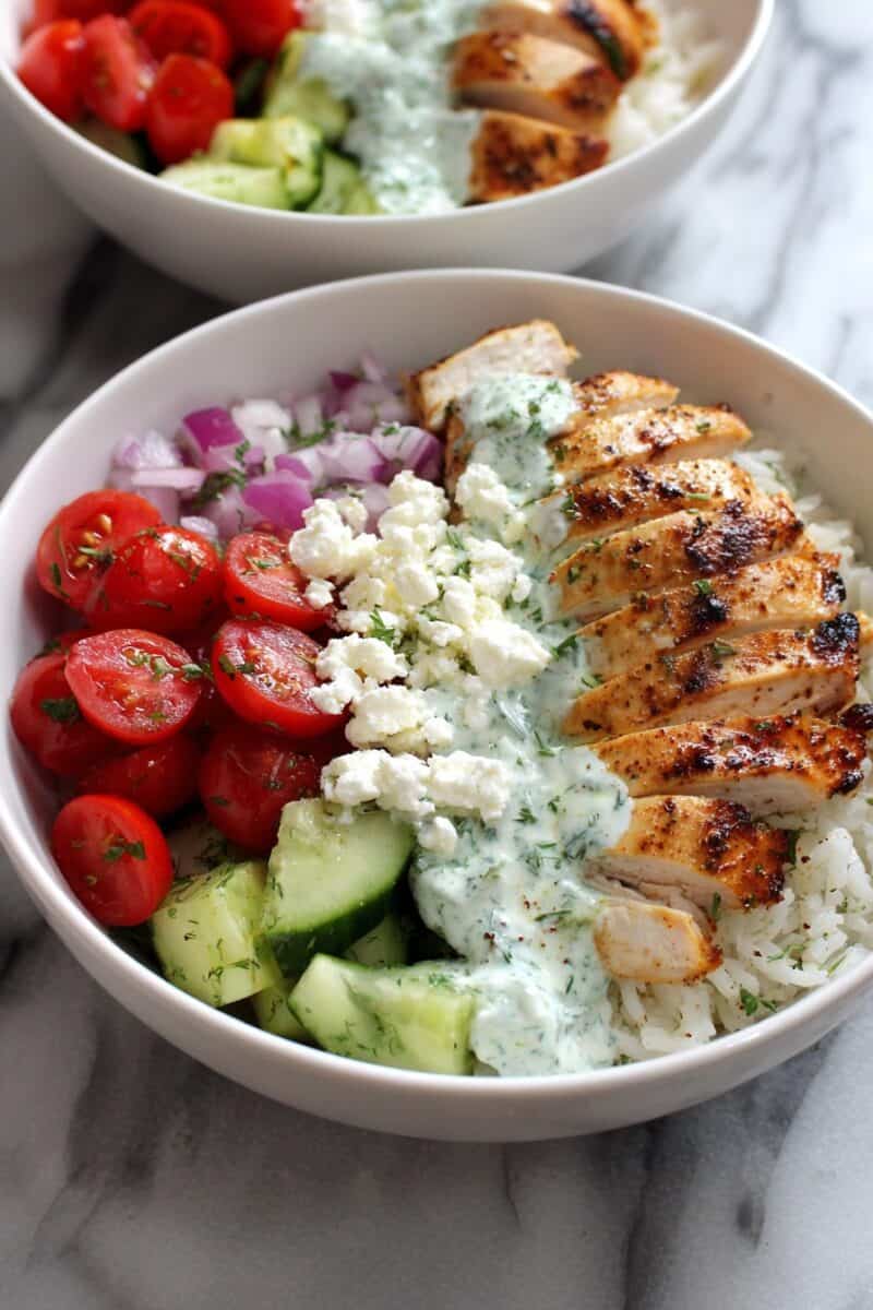 Clean eating greek chicken bowls topped with grilled chicken, rice, cucumber slices, cherry tomatoes, red onion, feta, and creamy greek yogurt sauce.
