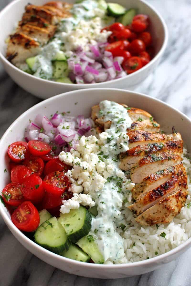 Healthy greek chicken bowls layered with lemon herb chicken, fluffy rice, fresh tomatoes, cucumbers, feta crumbles, and homemade tzatziki sauce in white serving bowls.
