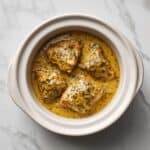 slow cooker filled with chicken thighs in a creamy herb sauce, shown from above in a white crockpot