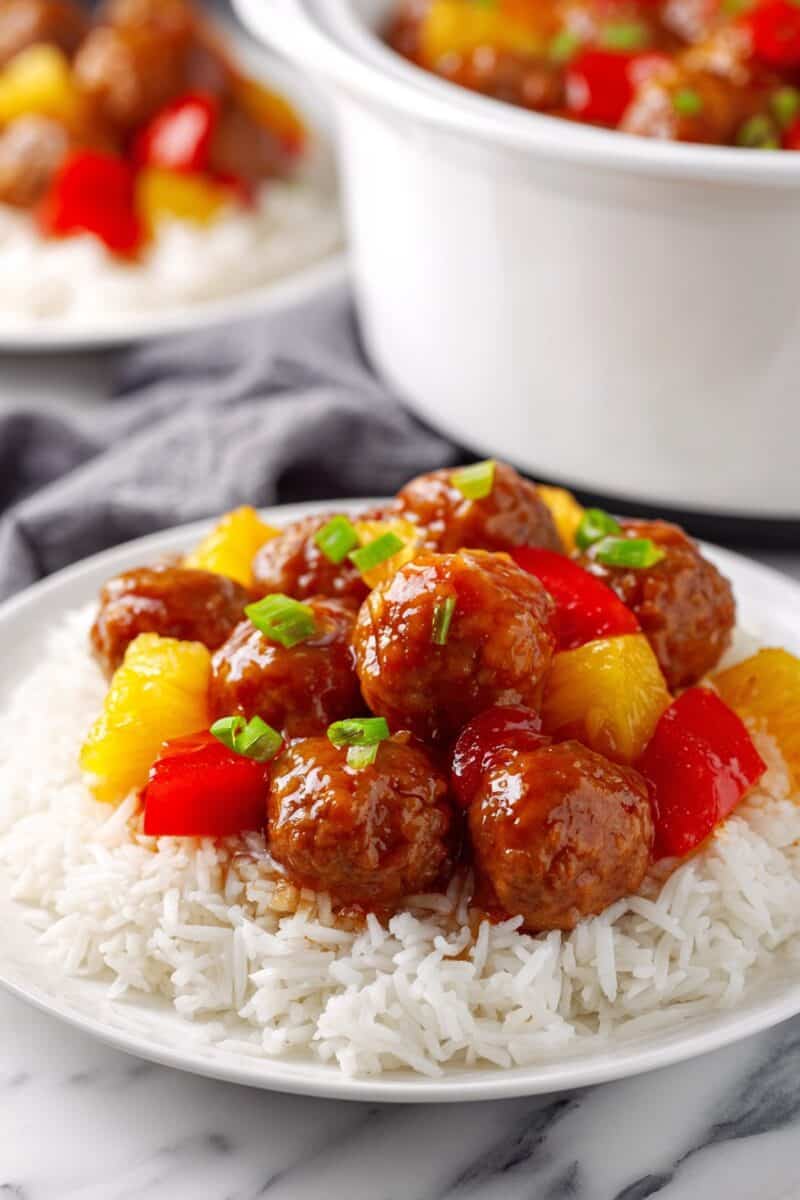 Sweet and sour chicken meatballs served over rice with pineapple and peppers with crockpot in background.