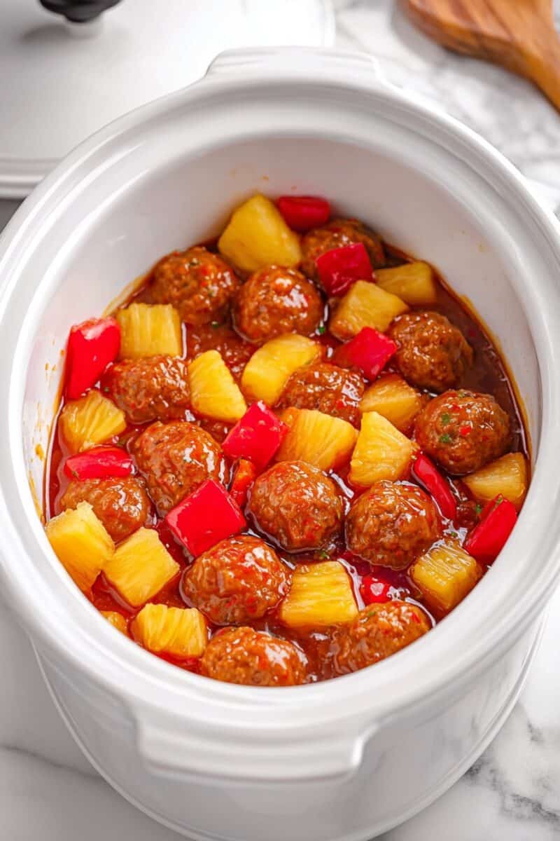 Sweet and sour chicken meatballs cooking in a crockpot with pineapple chunks and red bell peppers in thick sauce