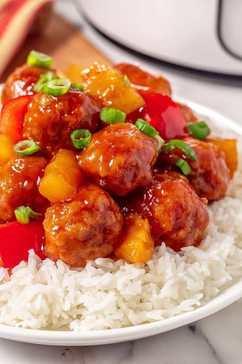 Close up of sweet and sour chicken meatballs coated in glossy sauce with pineapple and green onions over rice.