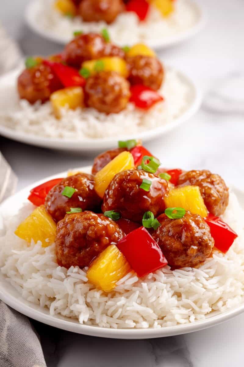 Sweet and sour chicken meatballs served over white rice with pineapple chunks and red bell peppers with extra plate in background