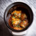 Close-up of tender chicken pieces cooking in a crockpot, coated in a glossy golden-brown sauce with visible herbs on top. The chicken appears juicy and fully cooked, sitting in a rich, slightly oily sauce inside a black slow cooker insert, perfect for an easy dump and go summer crockpot recipe.