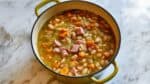 Close-up of high protein low calorie ham and bean soup with white beans, diced ham, carrots, celery, and herbs in a yellow Dutch oven.