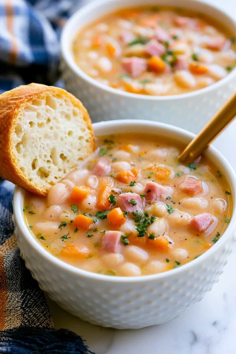 Bowl of ham and bean soup with white beans, diced ham, carrots, and herbs served with a slice of crusty bread.