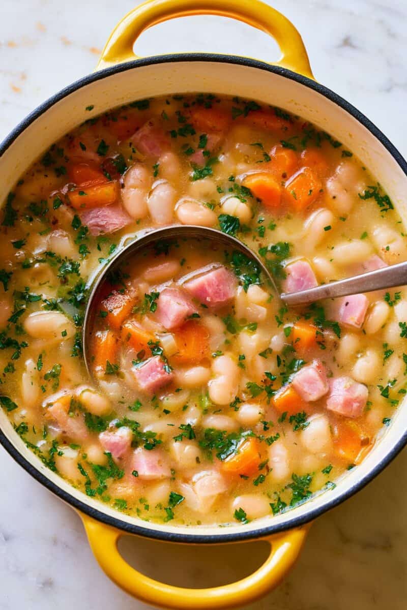 Overhead view of ham and bean soup with white beans, diced ham, carrots, and chopped herbs simmering in a yellow Dutch oven with a ladle.