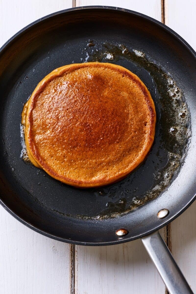 Single round pumpkin pancake cooking in the center of a black nonstick skillet, with a deep golden-brown surface and a small amount of melted cooking fat around the edges.