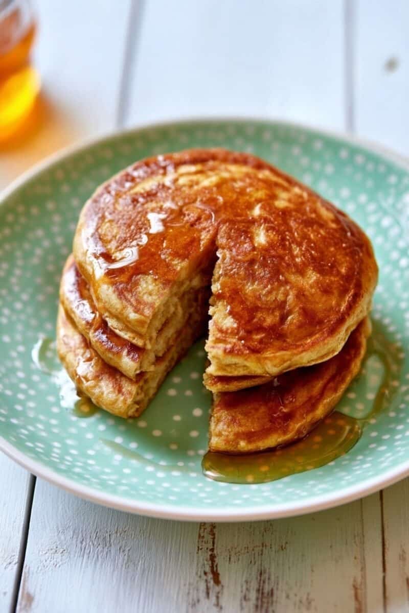 Three golden-brown pumpkin pancakes stacked on a green polka-dot plate with a wedge cut from the center, showing the thick, soft interior, with maple syrup drizzled over the pancakes and plate.