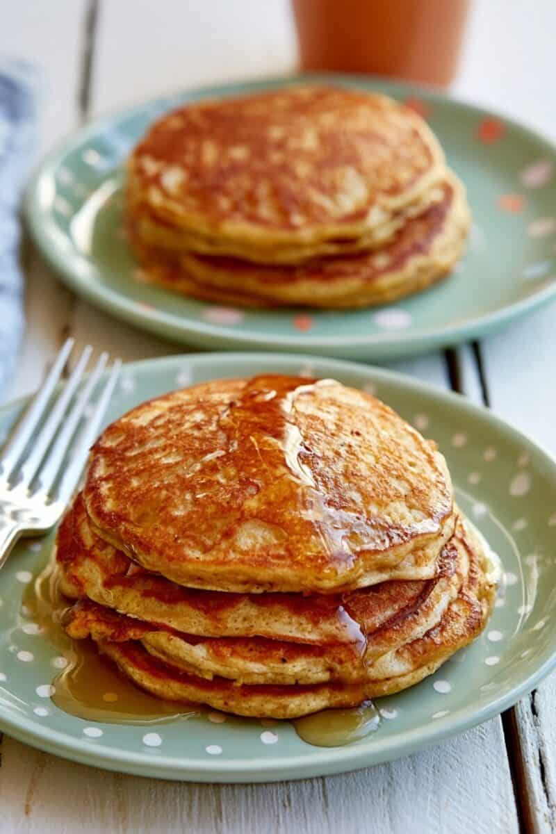 Stack of four thick golden-brown pumpkin pancakes on a green plate, topped with maple syrup and a small pat of butter, with a second plate of pancakes softly blurred in the background.