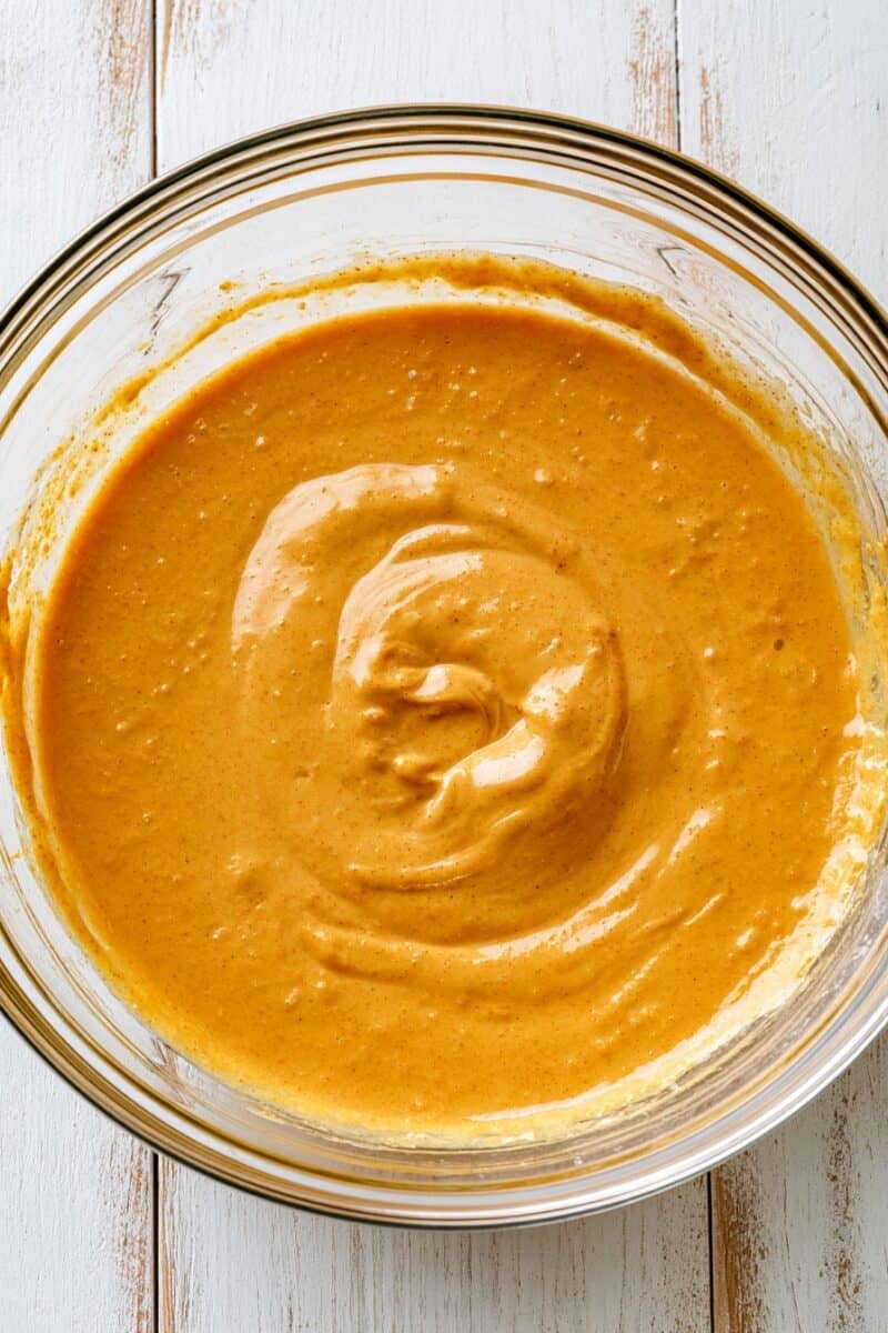Thick orange pumpkin pancake batter in a clear glass mixing bowl, with visible swirls from stirring and tiny specks of warm spices throughout the batter.