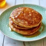 Stack of three thick golden-brown pumpkin pancakes on a green polka-dot plate with maple syrup being poured over the top pancake and dripping down the sides.