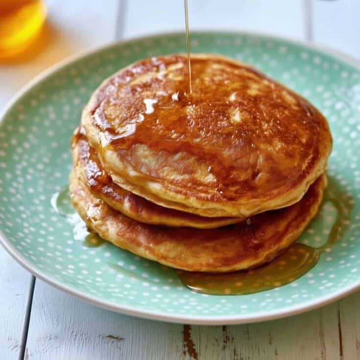 Stack of three thick golden-brown pumpkin pancakes on a green polka-dot plate with maple syrup being poured over the top pancake and dripping down the sides.