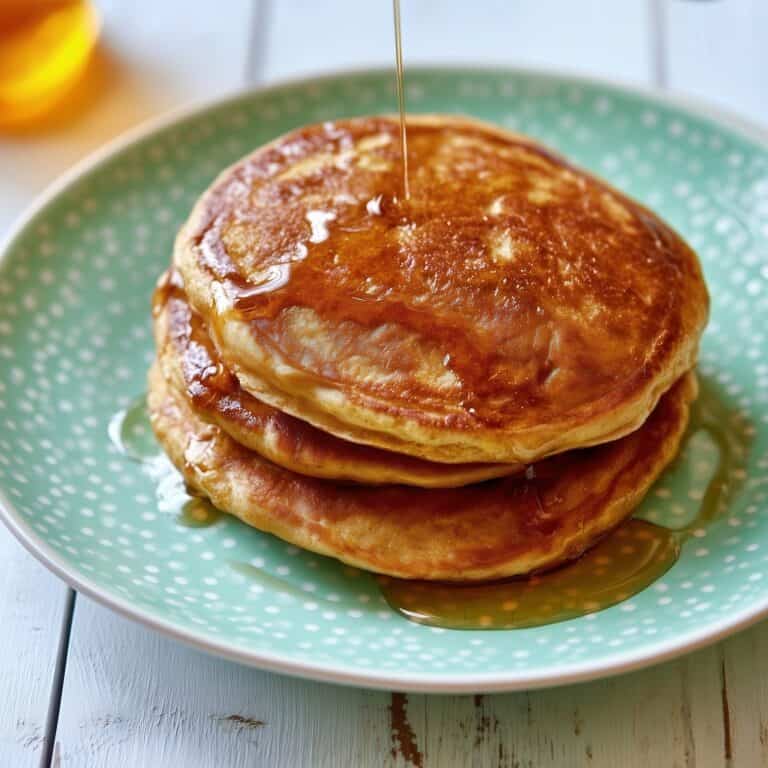 Stack of three thick golden-brown pumpkin pancakes on a green polka-dot plate with maple syrup being poured over the top pancake and dripping down the sides.